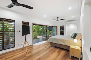 Ein modernes Schlafzimmer mit einem großen Bett, einem Fernseher auf einem Stativ, einem Deckenventilator und Blick auf einen Garten durch eine Schiebetür.