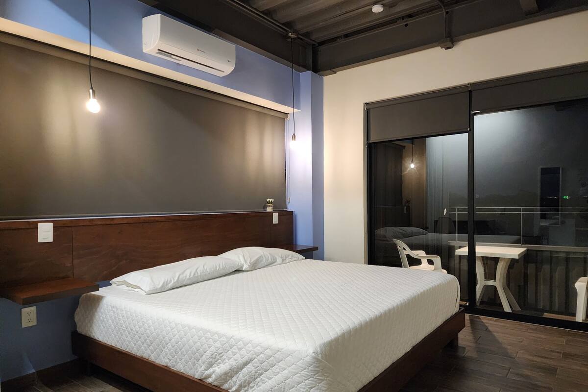 A modern hotel room with a wooden bed, a built-in bench, a small table, and a sliding glass door leading to a balcony.