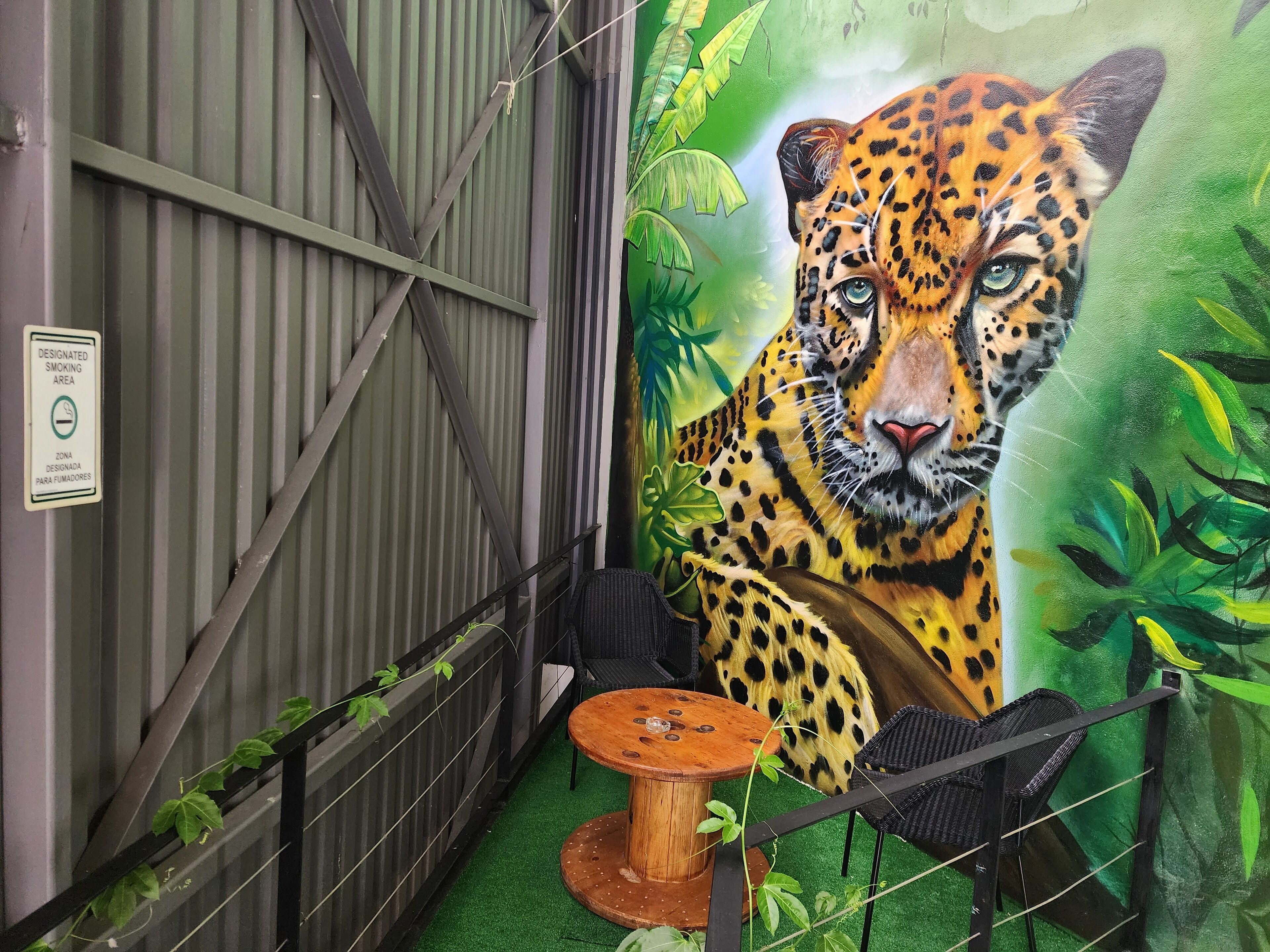 A room with a mural of a leopard, a designated smoking area sign, a small round wooden table, and a black wicker chair.