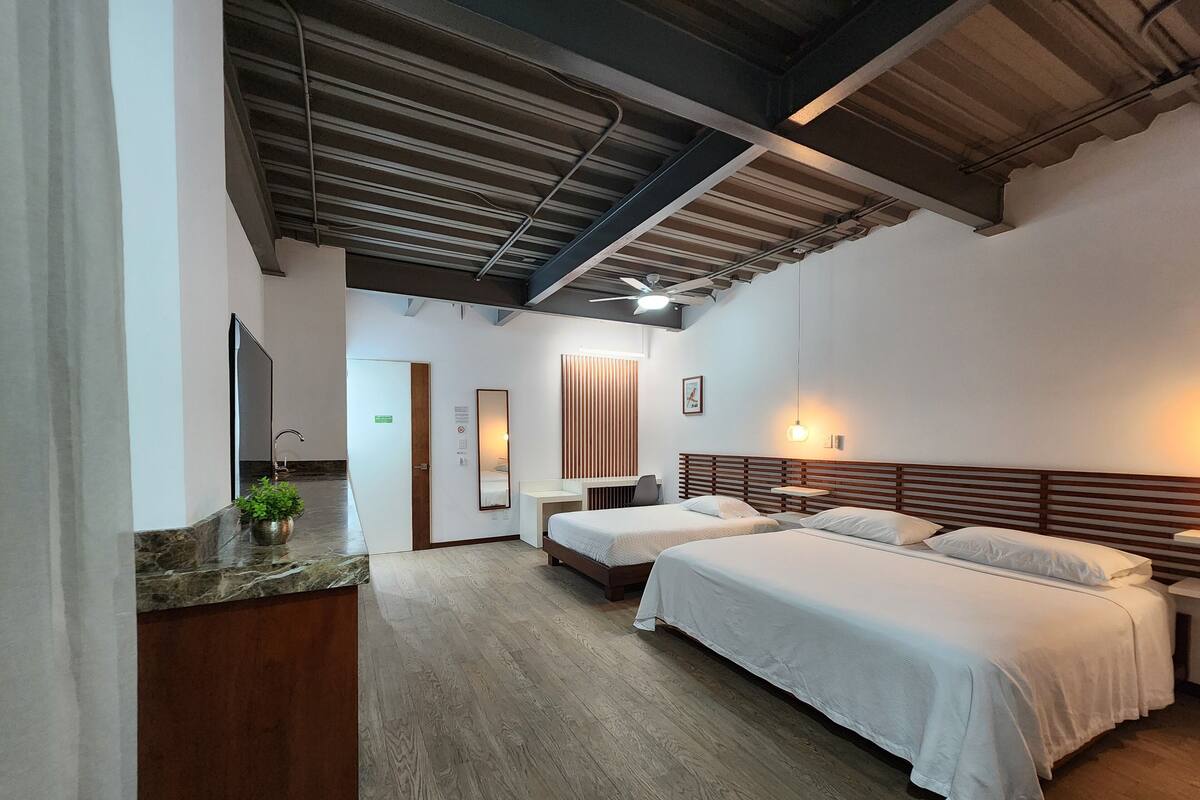 A hotel room with two beds, a wooden headboard, a small bedside table, a mirror, a potted plant, and a bathroom with a sink and mirror.
