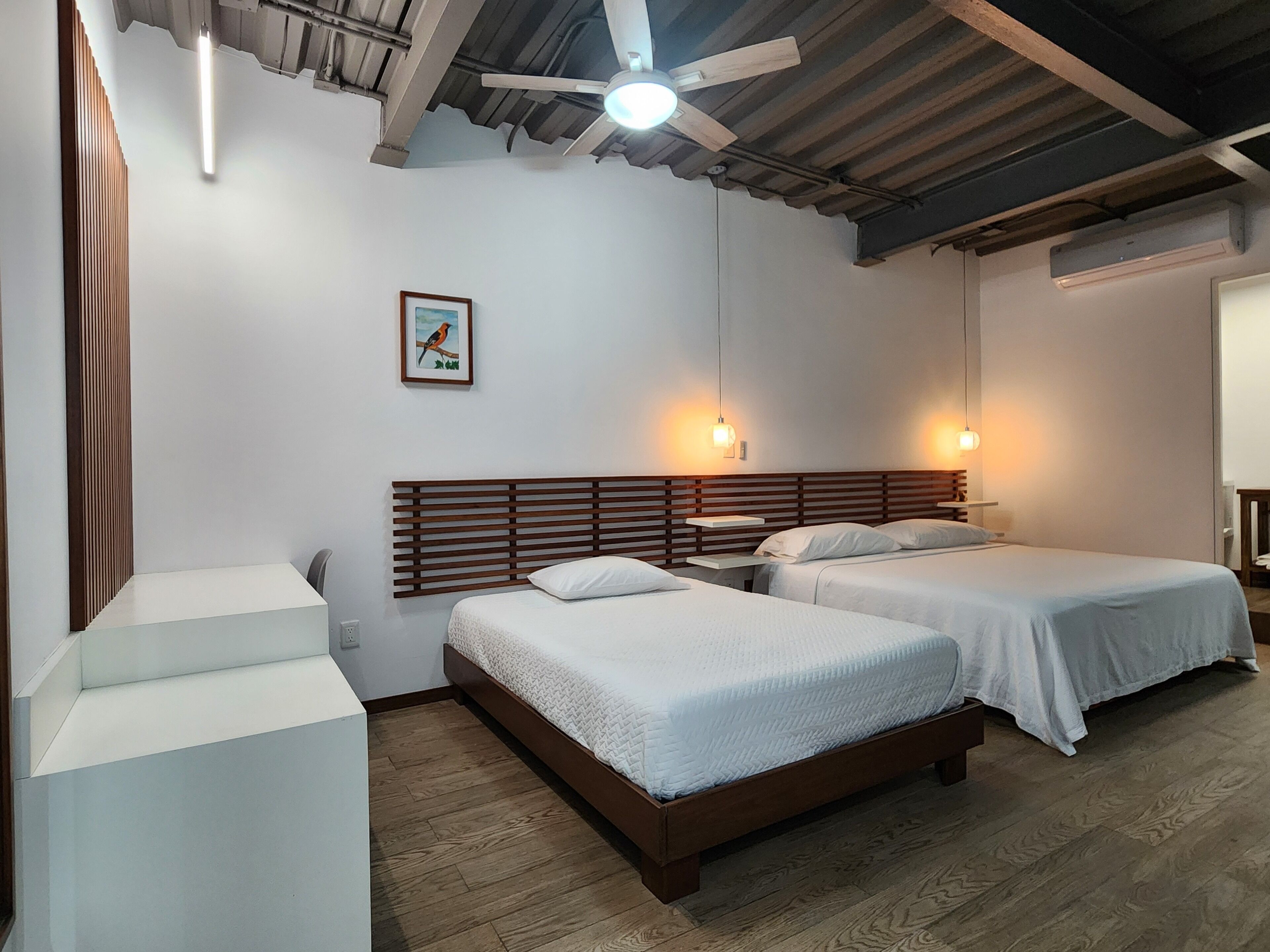 Habitación de hotel con dos camas, cabeceros de madera, un ventilador de techo y un cuadro en la pared.