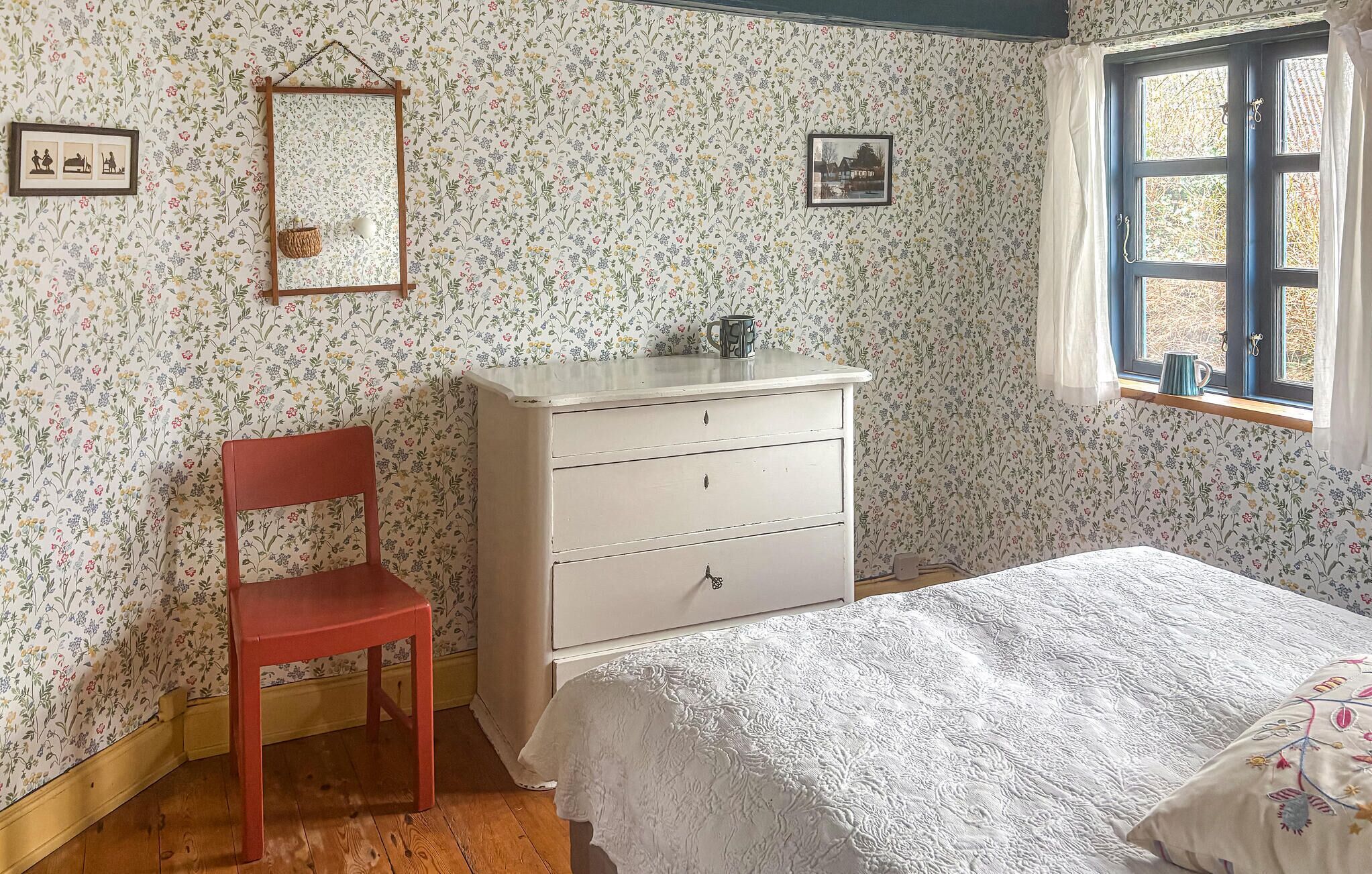 A bedroom with floral wallpaper, a wooden chair, a white dresser, a bed with a white bedspread, and a window with curtains.