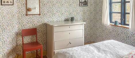 A bedroom with floral wallpaper, a wooden chair, a white dresser, a bed with a white bedspread, and a window with curtains.