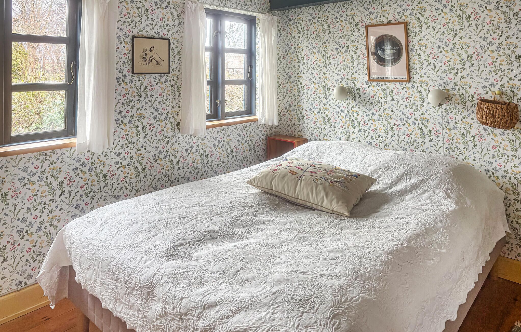Un dormitorio con papel tapiz floral, una cama con colcha de encaje blanco, un cuadro enmarcado en la pared y una ventana con cortinas.