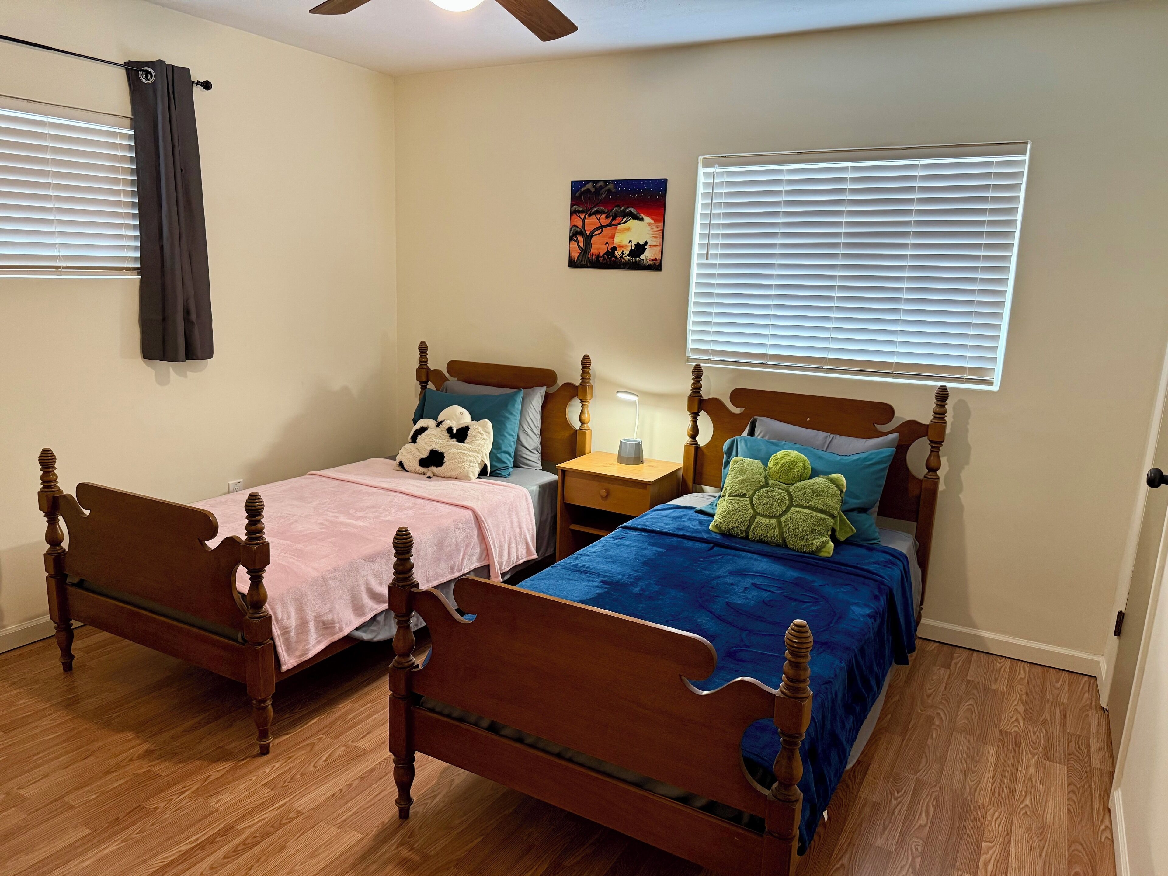A bedroom with two wooden beds, a nightstand, a ceiling fan, and a painting on the wall.