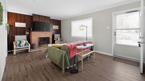 A cozy room with a brick fireplace, a bed with a green blanket, a small table, and a TV mounted on the wall. - Central STL - Near Zoo, WashU, Quiet w/ Easy Parking! (St. Louis)