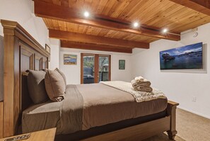 Une chambre avec un plafond en bois, un lit recouvert d’une couverture marron, un tableau au mur et une fenêtre donnant sur un paysage.