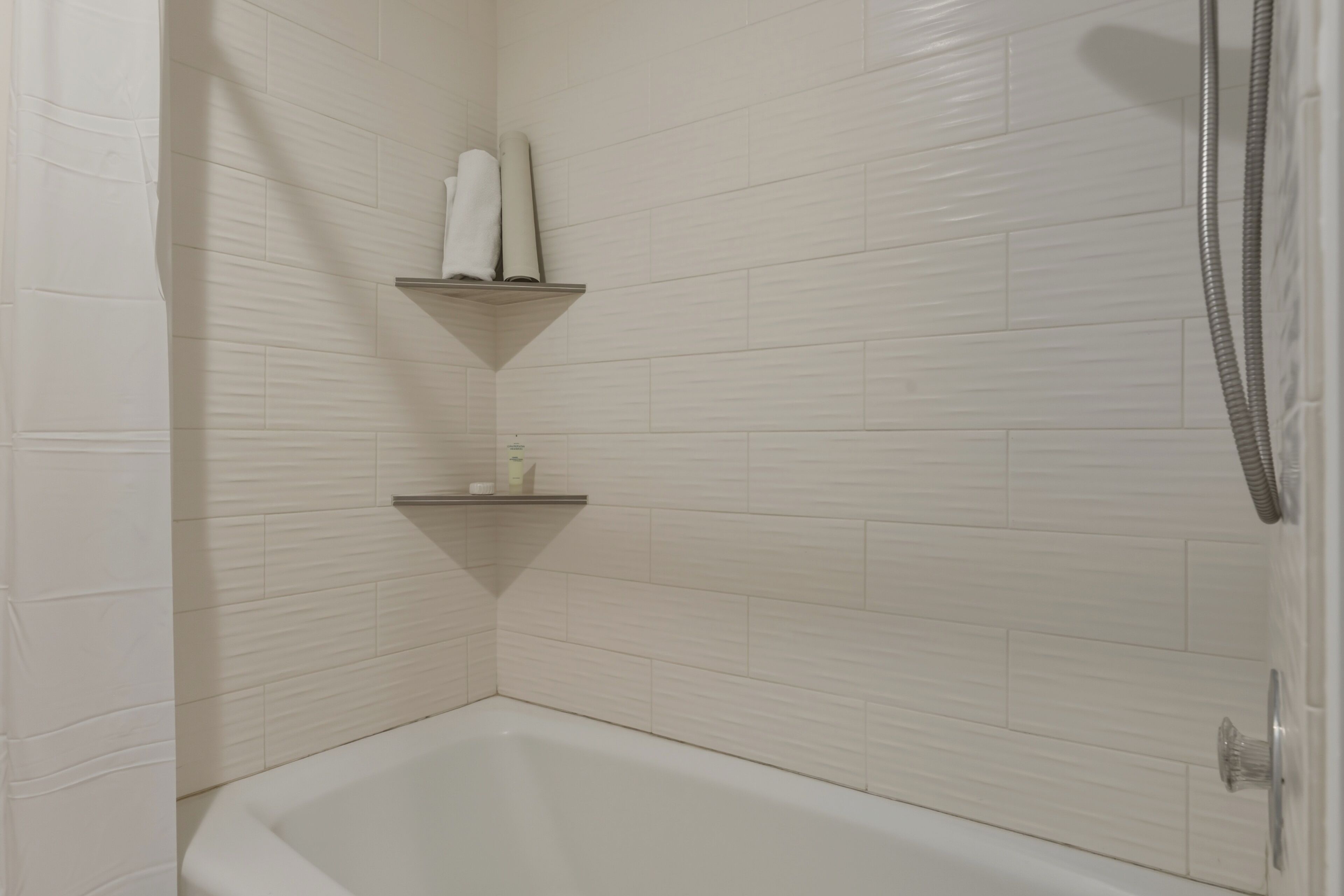 Ein Badezimmer im Eckbereich mit einer weißen Badewanne, zwei Ablagen mit Toilettenartikeln und einer Duschbrause.