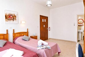 Une chambre d’hôtel avec deux lits, une tête de lit en bois, une table de chevet avec une lampe, un tableau au mur, une porte avec un détecteur d’incendie et une salle de bain visible à travers une porte ouverte.