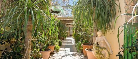 A narrow pathway flanked by potted plants and a statue, leading to a building with a green roof.