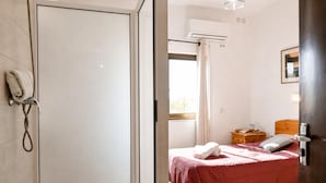 Habitación de hotel con ducha, cama con colcha roja, mesita de noche de madera y ventana con cortinas.