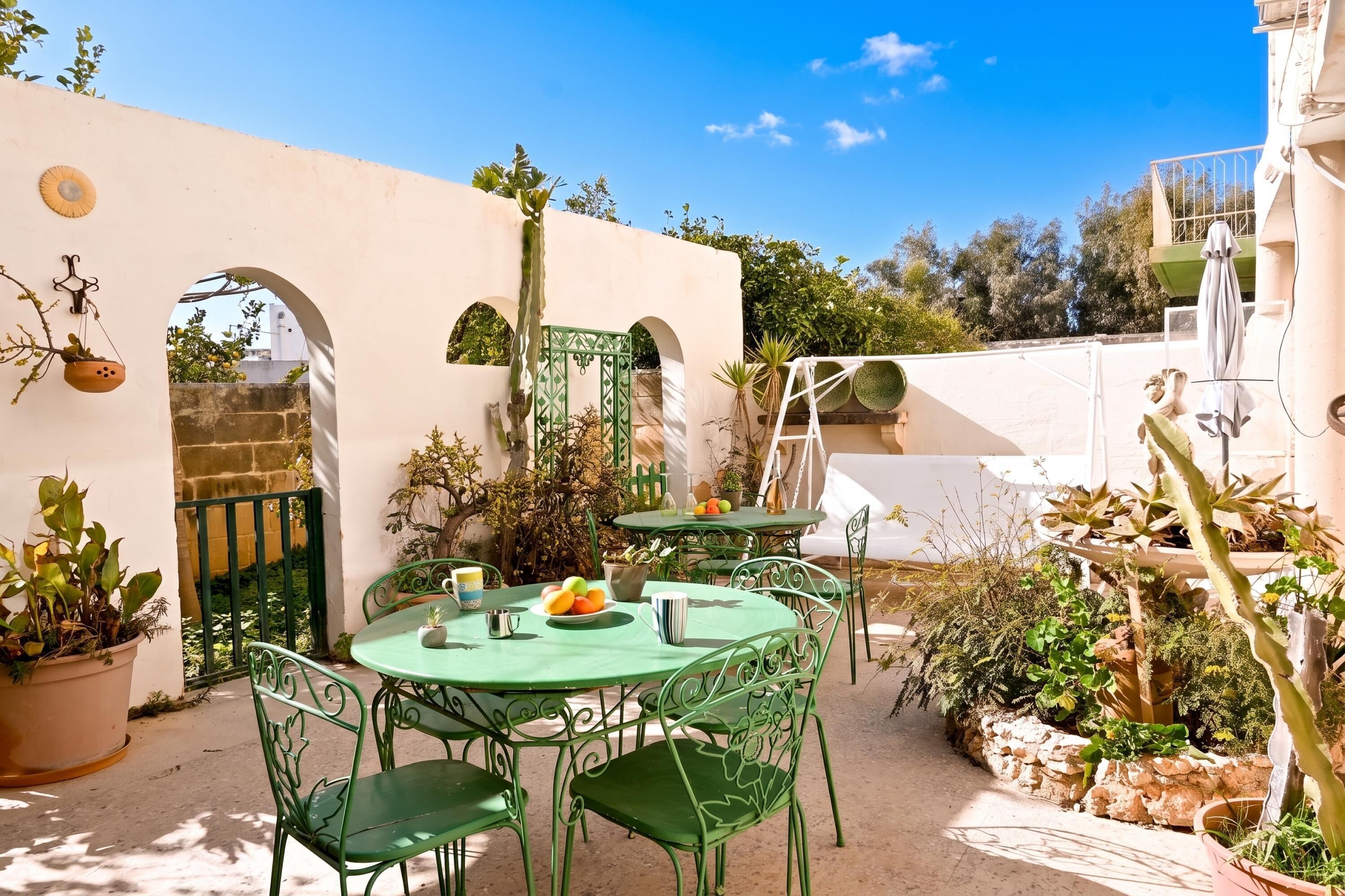 An outdoor patio with a round table and chairs, surrounded by potted plants and a white wall.