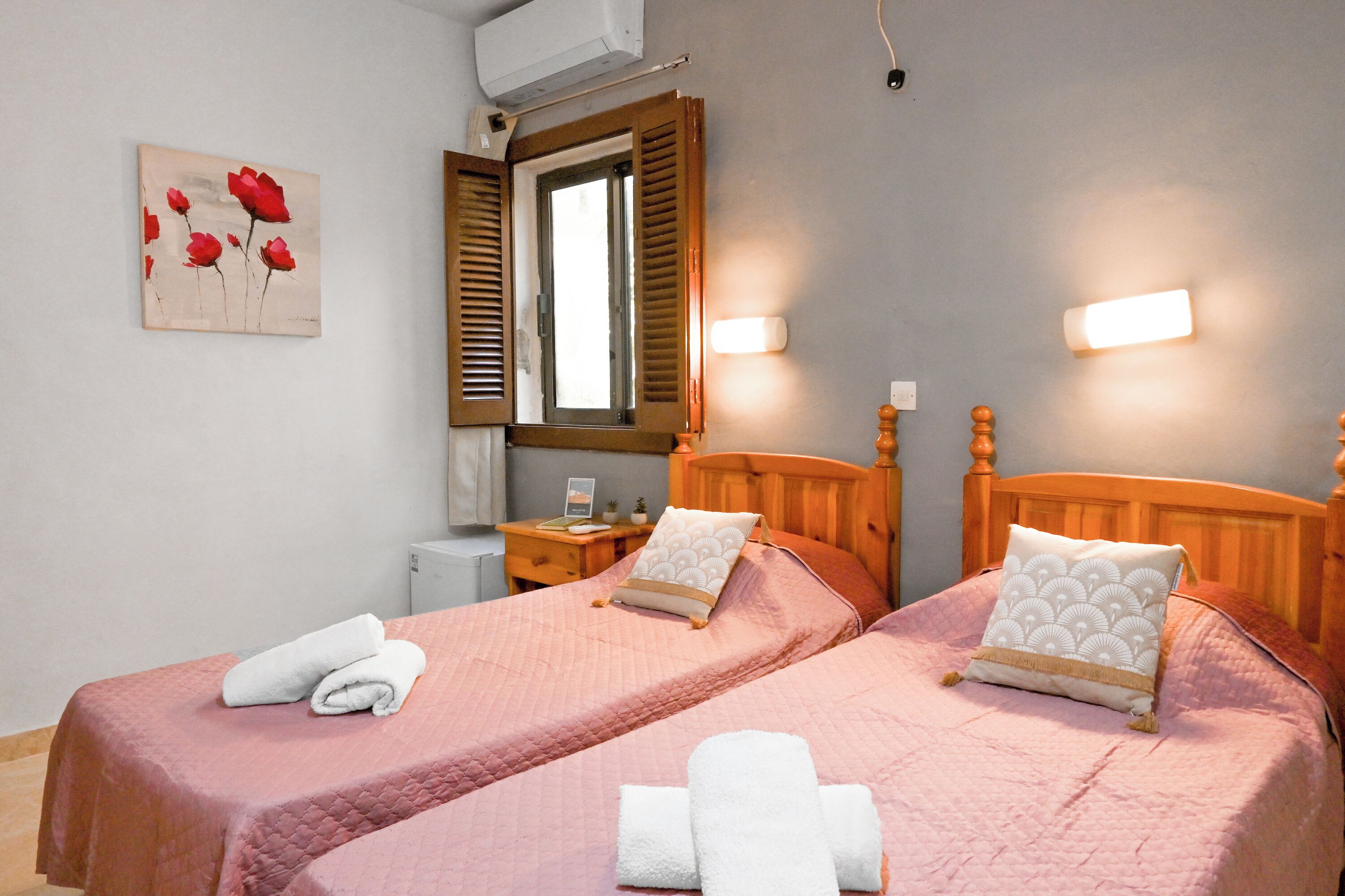 A hotel room with two beds, a window with wooden shutters, a bedside table with a lamp, and a wall-mounted air conditioner.