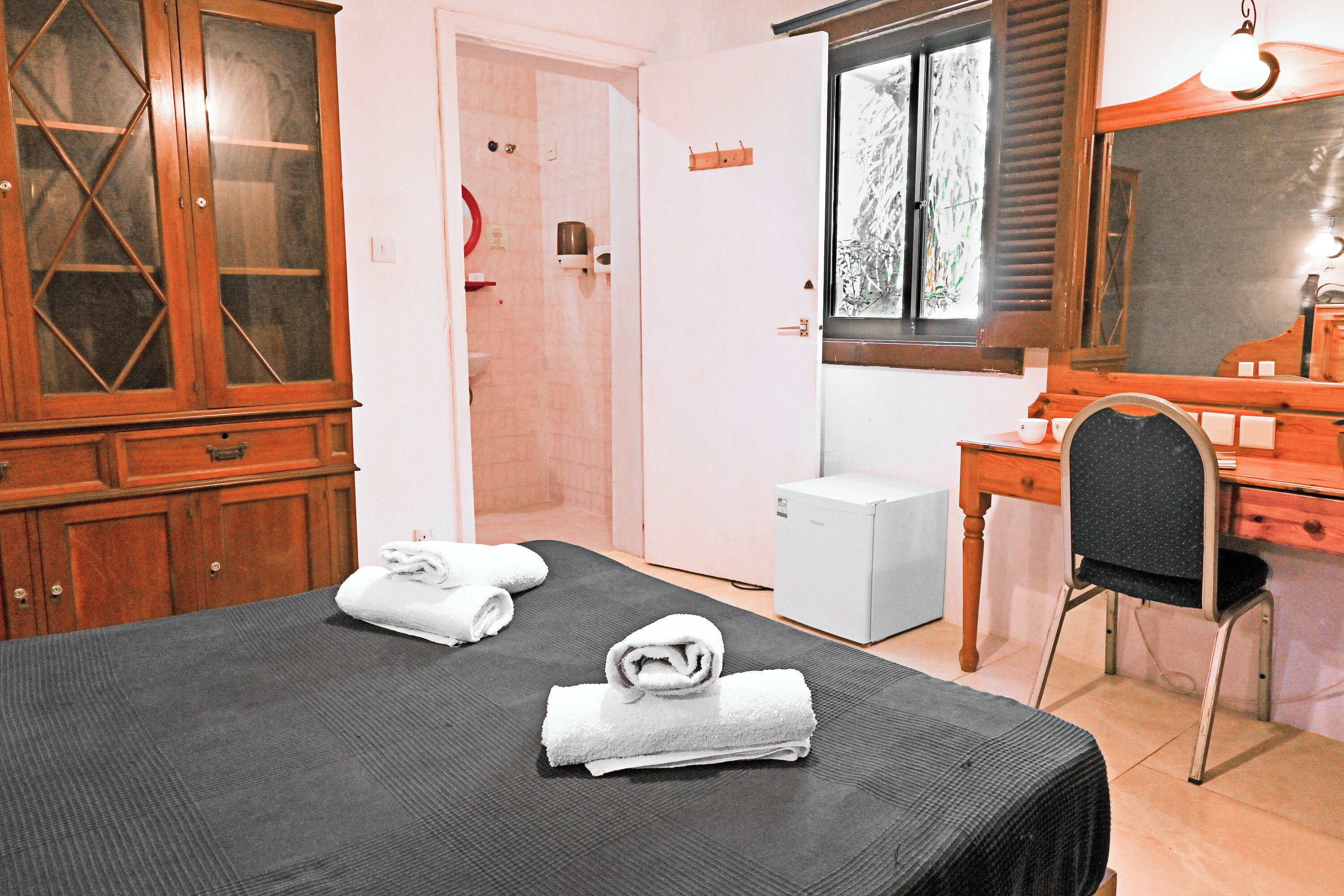 A hotel room with a bed, a desk, a chair, a wardrobe, and a small white appliance.