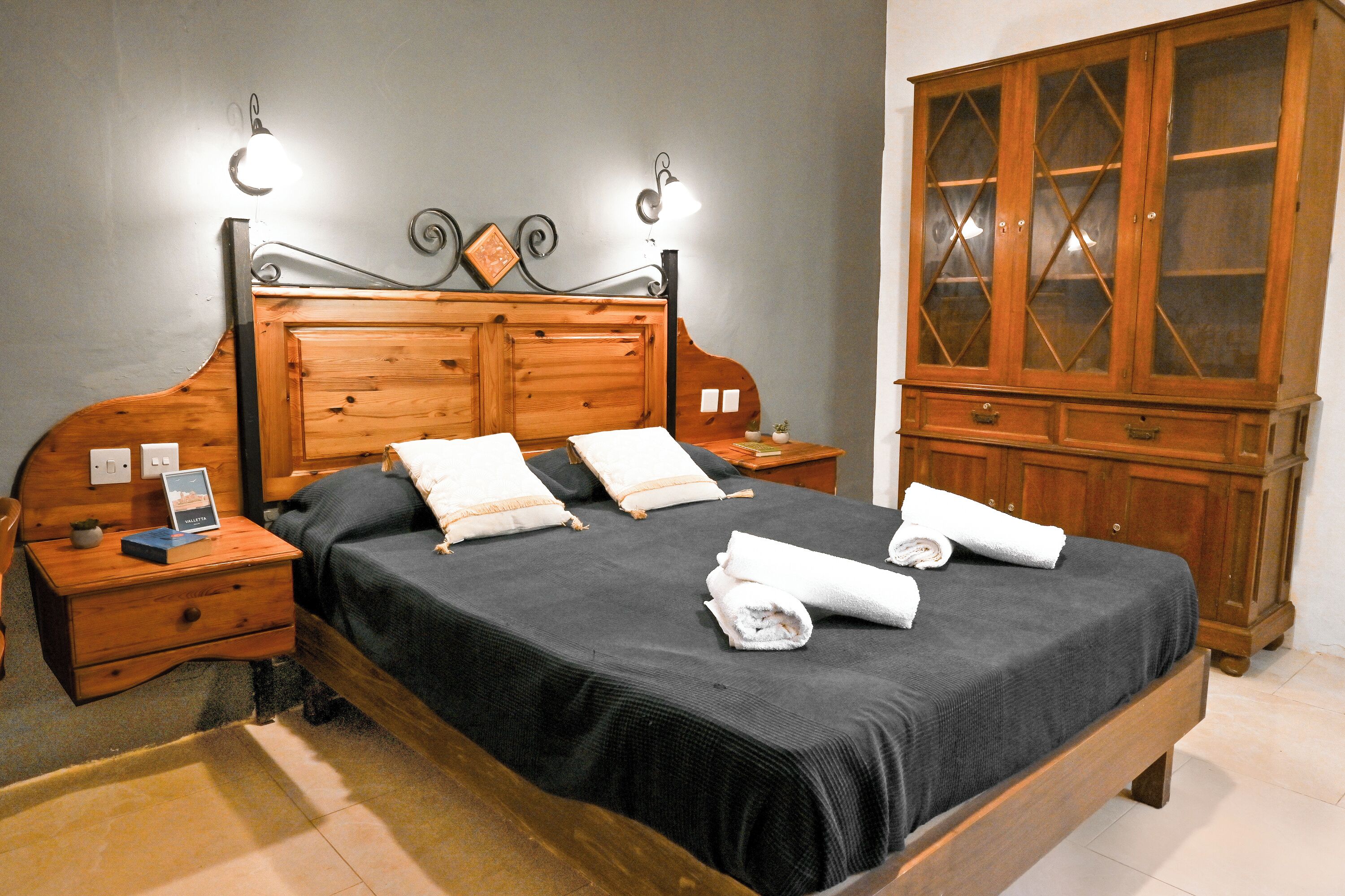 Ein ordentlich gemachtes Bett mit dunkler Bettdecke und Kissen, ein Nachttisch aus Holz mit einem Buch und einer kleinen Pflanze, ein Holzschrank mit Glastüren und eine Wandleuchte.