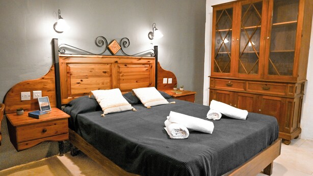 Un lit bien fait, avec une couverture sombre et des oreillers, une table de chevet en bois sur laquelle se trouvent un livre et une petite plante, une armoire en bois avec des portes en verre, et un luminaire fixé au mur.