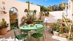Une terrasse extérieure avec une table ronde et des chaises, entourée de plantes en pot et d’un mur blanc.