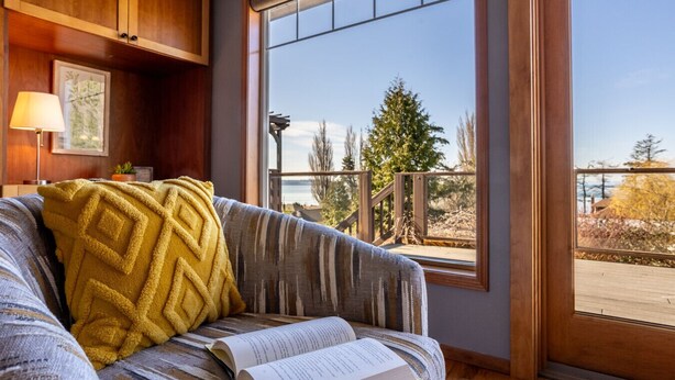 Ein gemütliches Zimmer mit großem Fenster, einem Sofa mit gemustertem Überwurf und Blick auf eine Holzterrasse und Bäume.