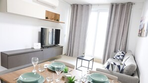 A modern living room with a dining area, grey curtains, a flat-screen TV, and a grey sofa with patterned cushions. - Central Sliema Stay Sea View -steps to Promenade (Sliema)