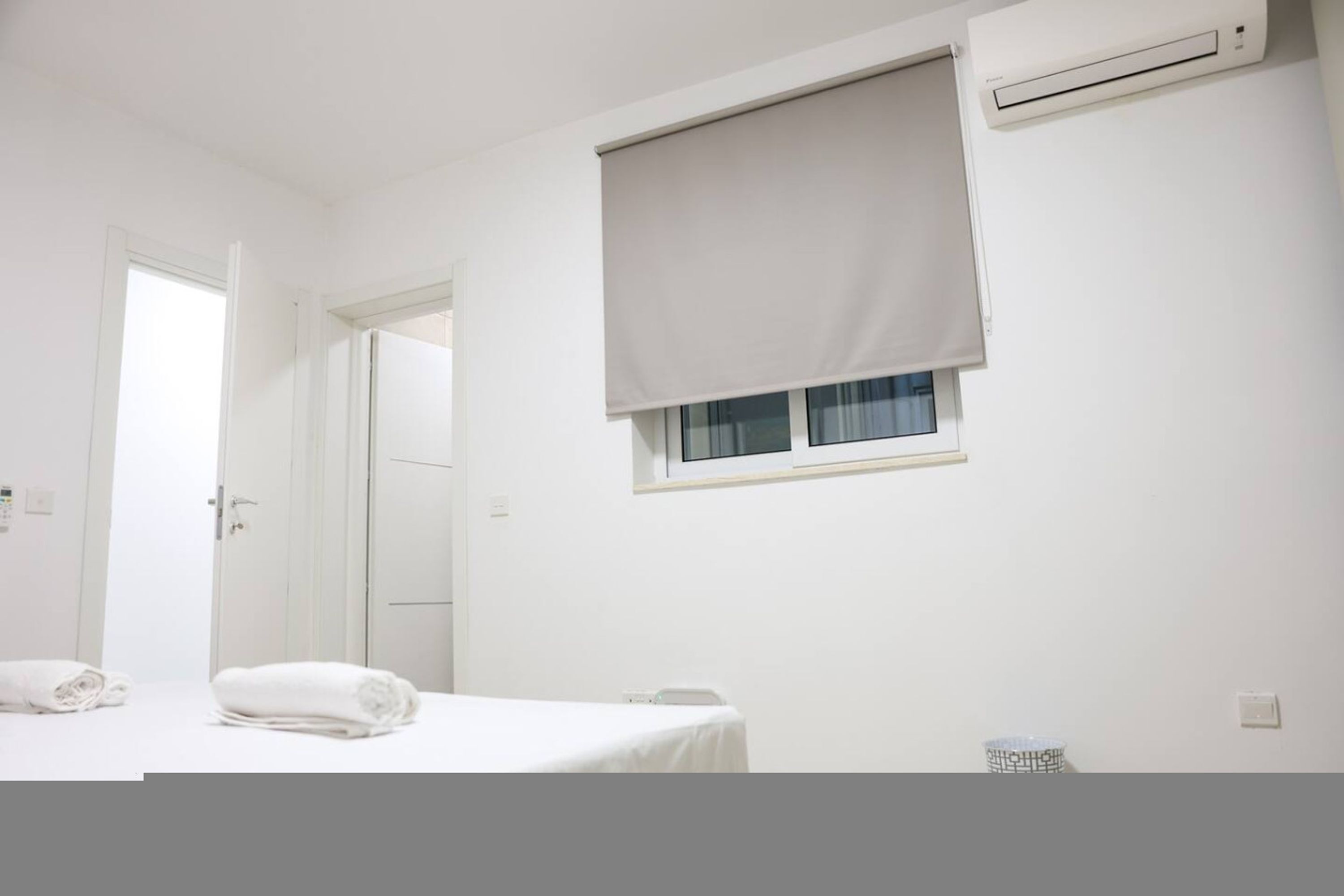 A hotel room with a bed, a window with a blind, an air conditioning unit, and a door.