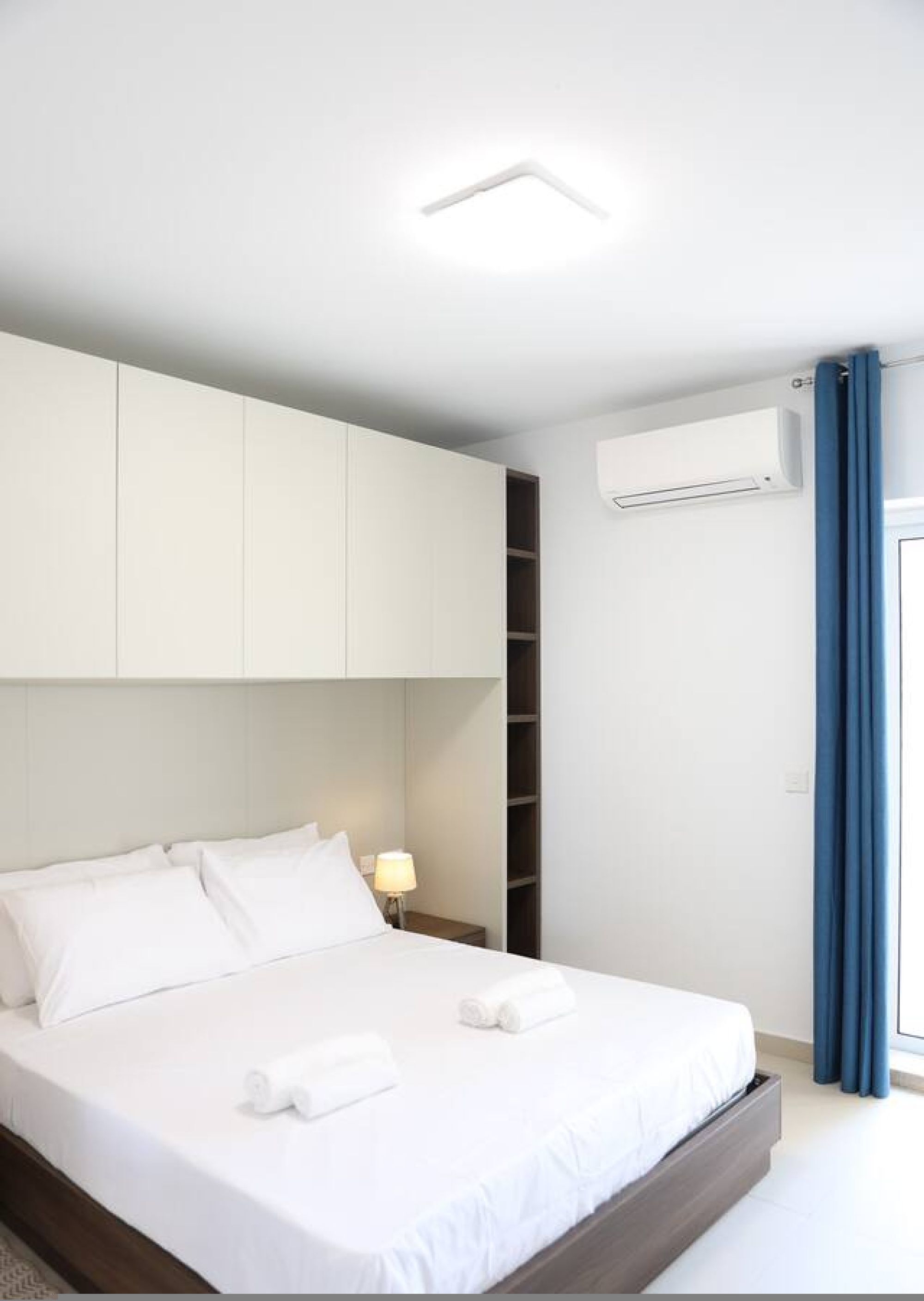 A modern hotel room with a bed, white linens, a nightstand, a lamp, a wardrobe, and an air conditioning unit.