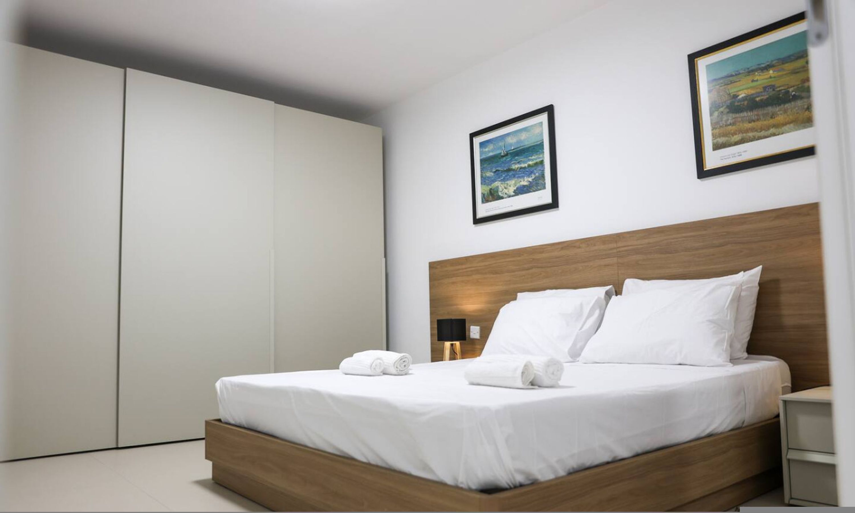 Een moderne hotelkamer met een houten hoofdeinde, witte beddengoed en twee ingelijste landschapsschilderijen aan de muur.