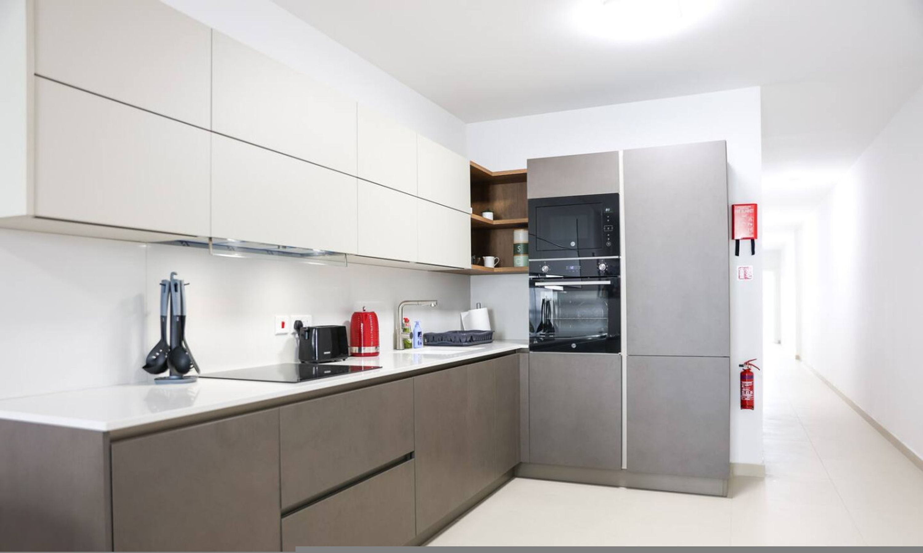 A modern kitchen with grey cabinets, a stainless steel refrigerator, a built-in oven, and a red fire extinguisher.
