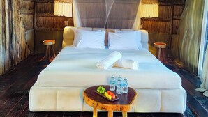 Un lit à baldaquin, une table de chevet avec des fruits et des bouteilles d’eau, et un plafond en chaume.