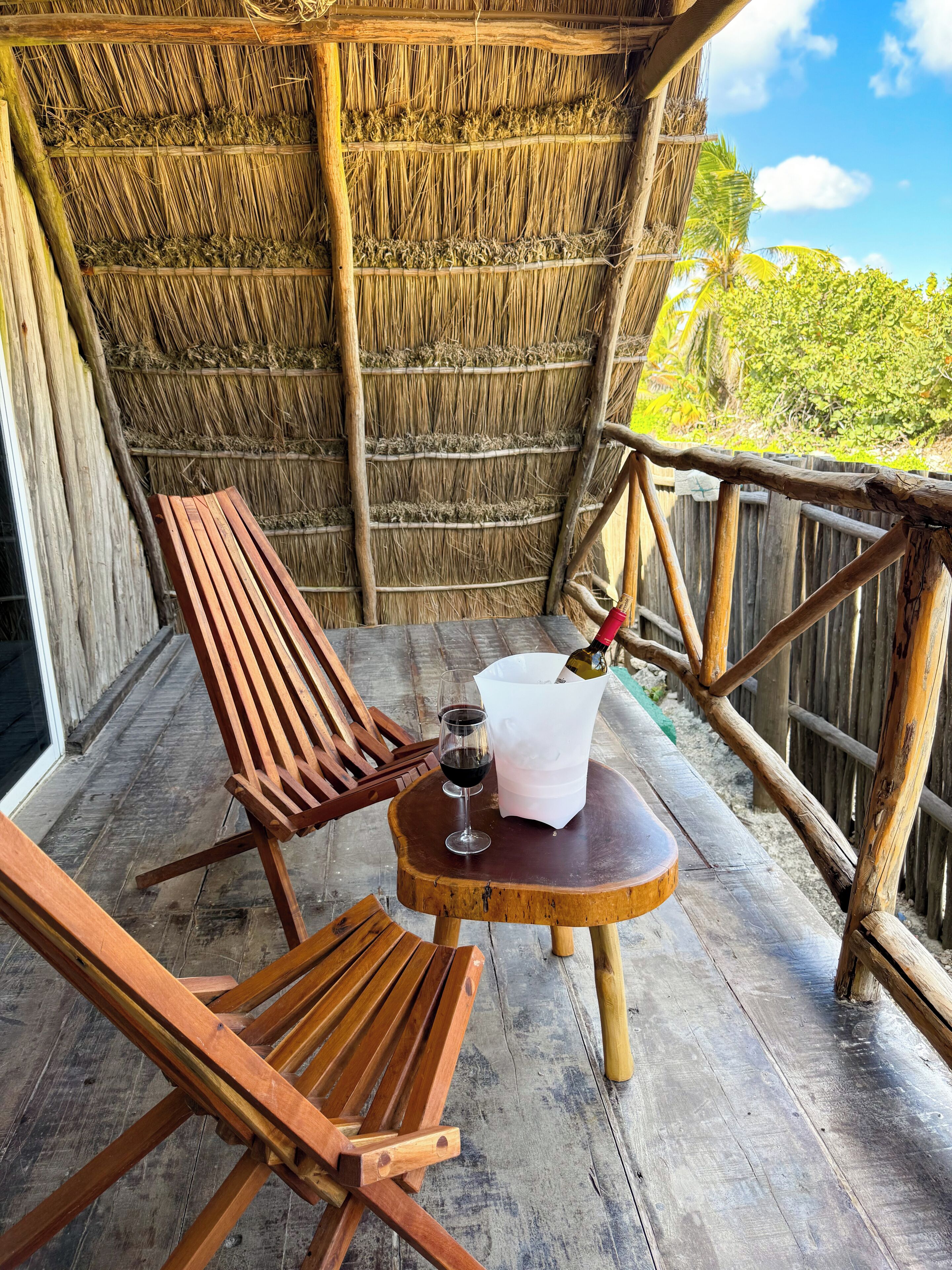 Une chaise et une table en bois sur un balcon doté d’un toit de chaume, une bouteille de vin et un verre.