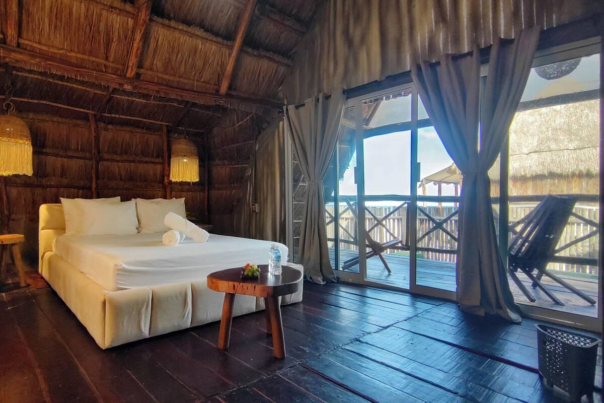 A wooden room with a bed, a small table, and a window with a view of a wooden deck.