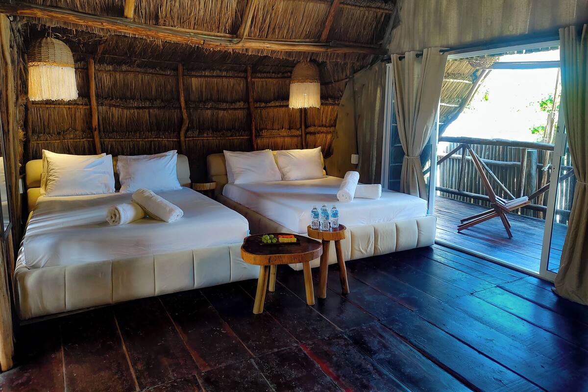 A room with two beds, a small table with bottles, and a view of the outside.