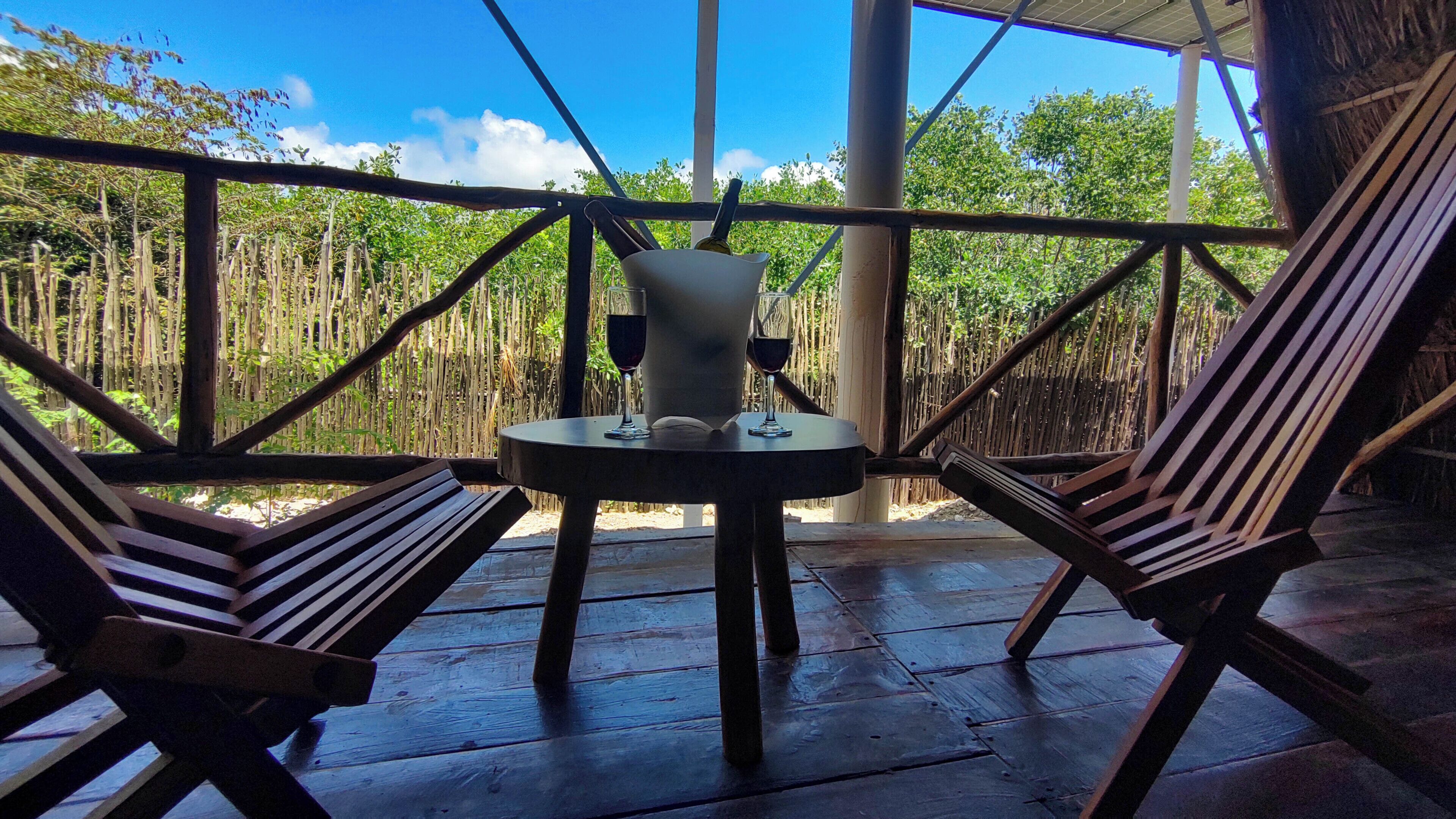 A wooden table with two chairs, a bottle, and two glasses on a veranda with a bamboo fence and trees in the background.