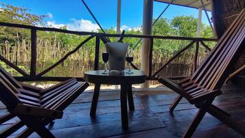 A wooden table with two chairs, a bottle, and two glasses on a veranda with a bamboo fence and trees in the background.