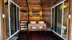 Une pièce avec un plafond en chaume, un banc en bois avec des serviettes, un lavabo avec un miroir et des toilettes.