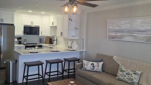 Une cuisine moderne avec des meubles blancs, un îlot central doté de tabourets, un ventilateur de plafond et un coin repas comprenant un canapé et une table basse.