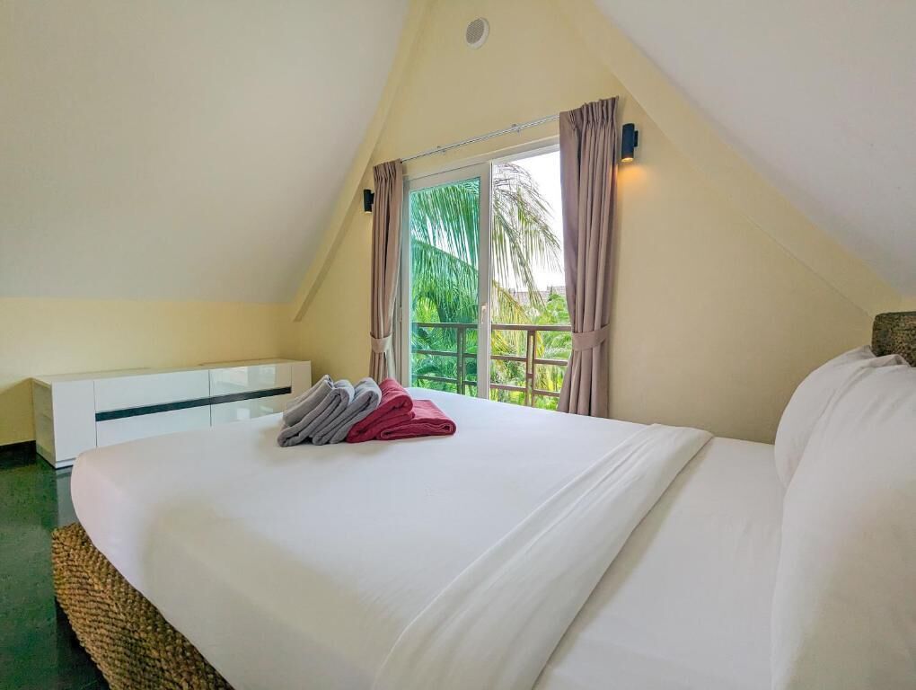 A bedroom with a large bed, a bedside table, and a view of greenery through a window.