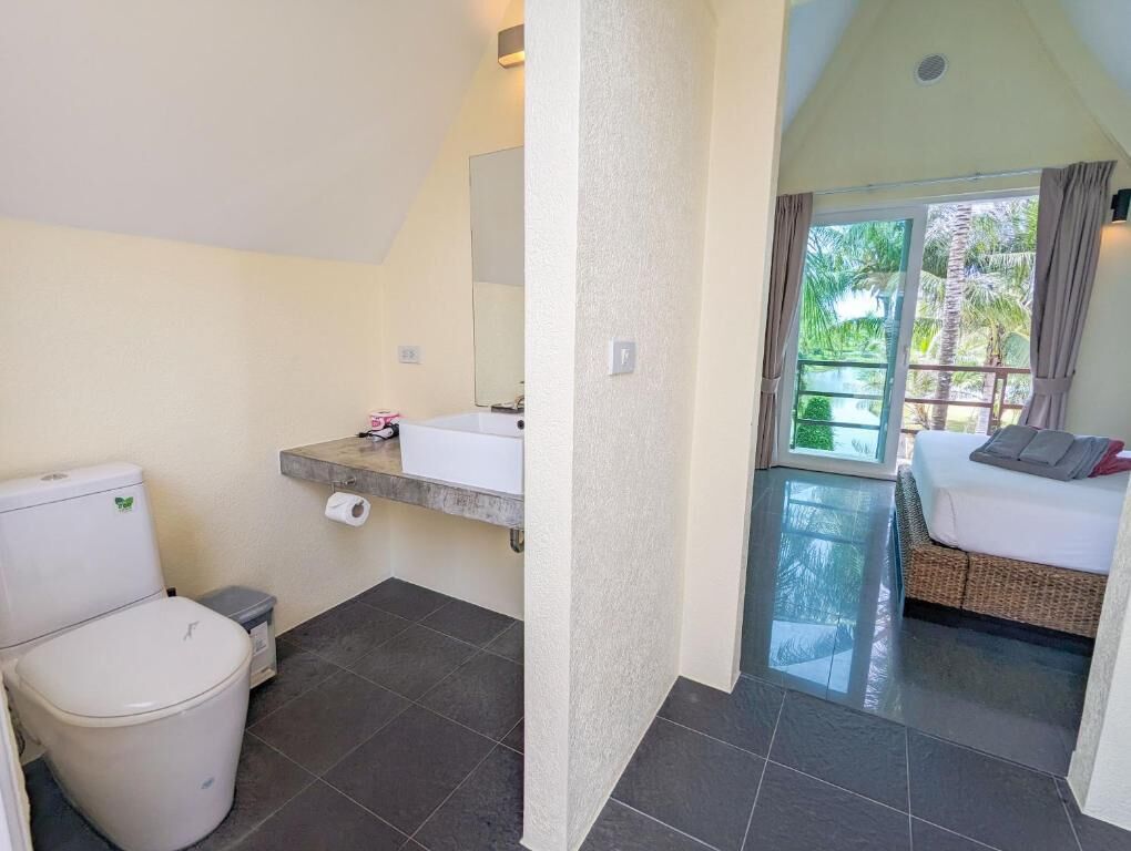 A bathroom with a toilet, sink, and shower area. There is a bed with a view of palm trees outside.