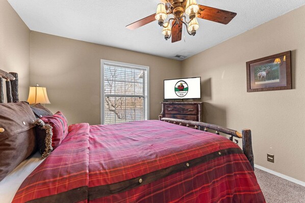 Een slaapkamer met een bed, een plafondventilator, een televisie, een raam met zonwering, en een ingelijste foto aan de muur.