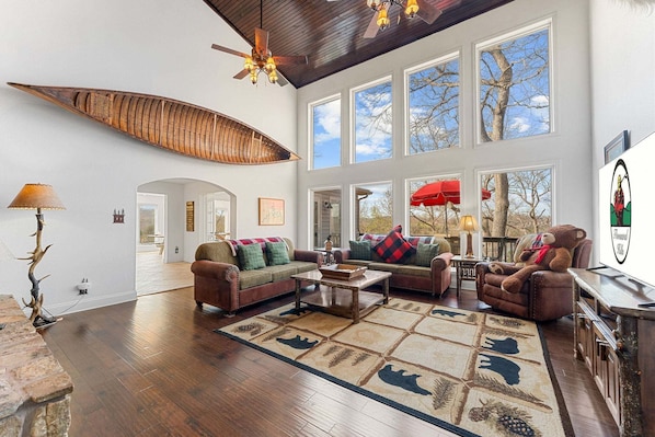 A spacious living room with a large wooden ceiling, a fireplace, a large window, and a fireplace mantel.