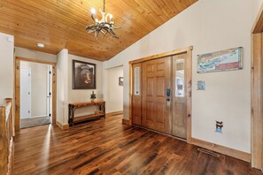 Un couloir au sol en bois, orné d’un lustre, avec une porte en bois et un tableau encadré accroché au mur.