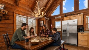 Interieur van een houten chalet met een eettafel, stoelen, een bank, een koelkast en een bordje met de tekst 'CABINS'.