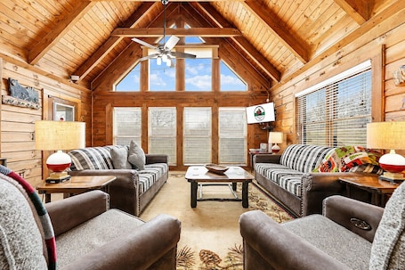 A log cabin interior with two sofas, a coffee table, a ceiling fan, and a television.