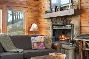 A cozy cabin interior with a stone fireplace, wooden walls, a leather sofa, and a window overlooking a wooden deck. - Thunder Ridge + Table Rock • Hot Tub • Fenced Yard • Pet Friendly (Ridgedale)