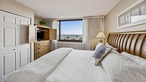 A hotel room with a large bed, a dresser, a desk with a computer, a window with a view of the sea, and a lamp. - Oceanview 3BR Condo - Beautiful Pool on Amelia Island (Fernandina Beach)