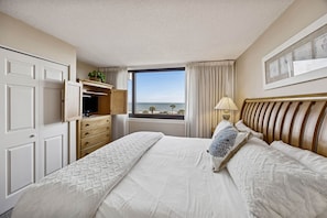 A hotel room with a large bed, a dresser, a desk with a computer, a window with a view of the sea, and a lamp. - Oceanview 3BR Condo - Beautiful Pool on Amelia Island (Fernandina Beach)