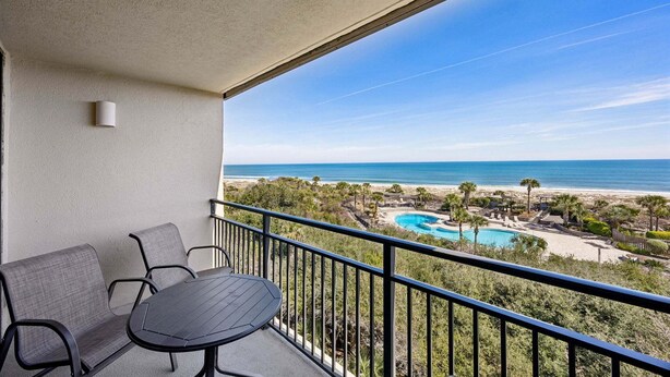 A balcony with a table and chairs, overlooking a beach and a swimming pool. - Oceanview 3BR Condo - Beautiful Pool on Amelia Island (Fernandina Beach)