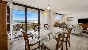 A spacious living area with a glass dining table, wooden chairs, a sofa, and a TV. Large windows offer a view of the ocean. - Oceanview 3BR Condo - Beautiful Pool on Amelia Island (Fernandina Beach)