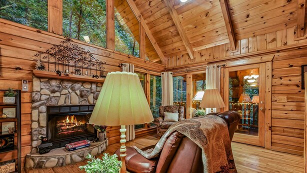 A log cabin interior with a stone fireplace, leather seating, and a large window overlooking the forest. - Mountain Joy: Free Tickets/ Jacuzzi/ Fireplace (Gatlinburg)