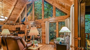 A cozy cabin interior with a fireplace, wooden furniture, and a large window overlooking the forest. - Mountain Joy: Free Tickets/ Jacuzzi/ Fireplace (Gatlinburg)