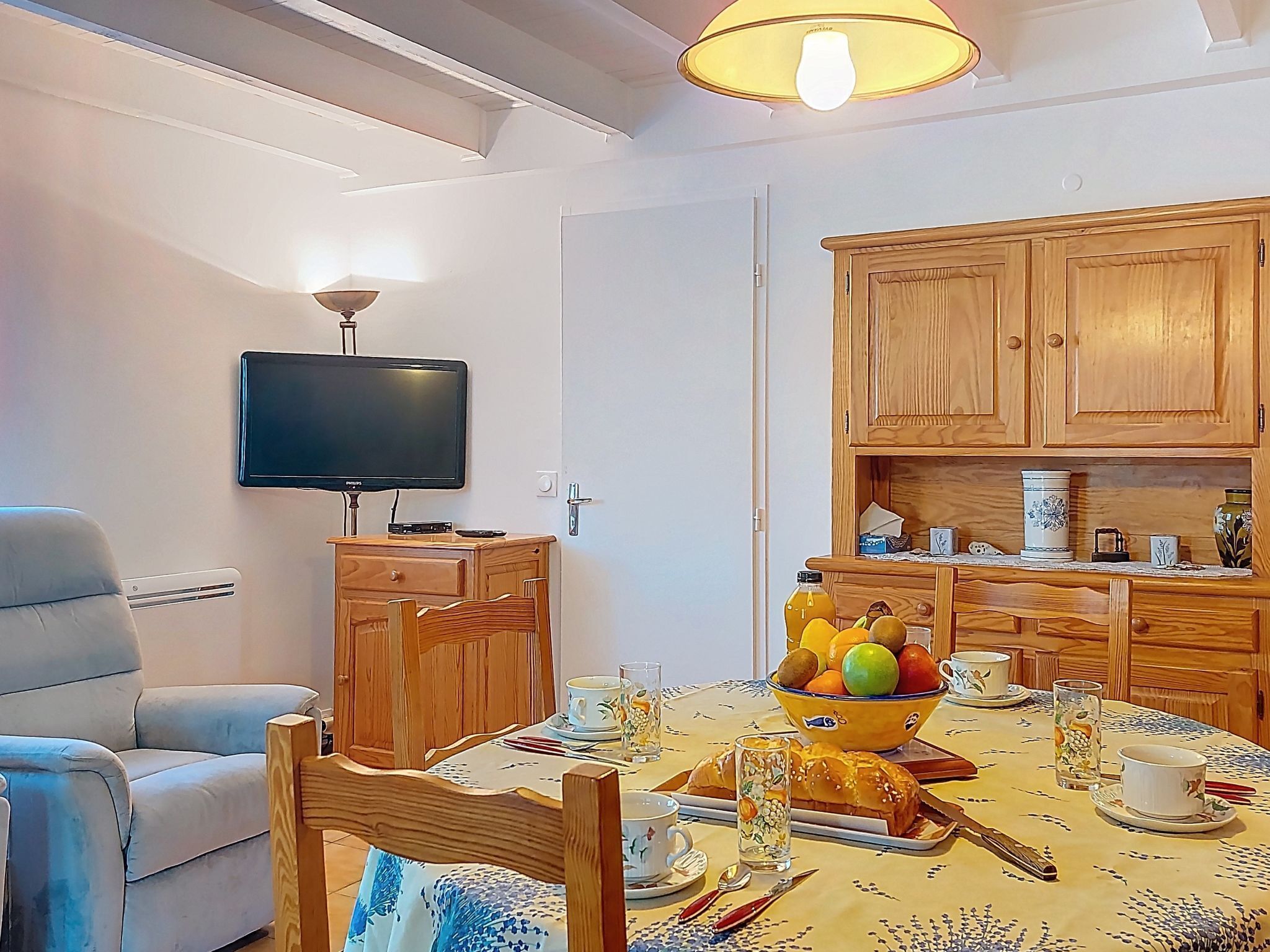 A dining area with a table set for two, a television mounted on the wall, wooden cabinets, and a ceiling light fixture.